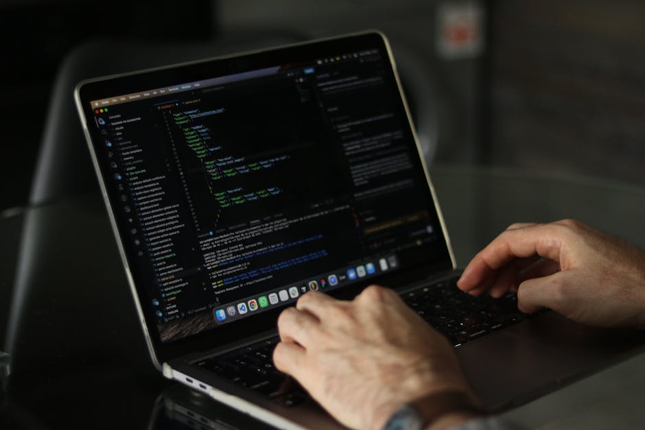 A close-up view of a person's hands typing on a MacBook laptop, which displays lines of code in a dark-themed programming environment. The individual appears focused, with the background slightly blur